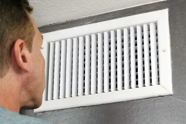 A person closely inspecting a white air vent mounted on a gray wall.